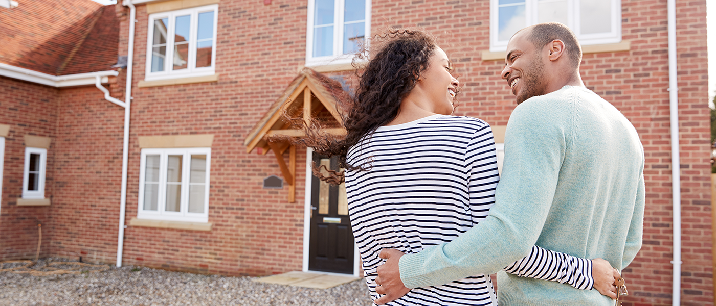 A couple smiling at each other outside a house for sale. Image represents a couple looking for a new home and possibly requiring a home mover mortgage with the help of an independent mortgage advisor.